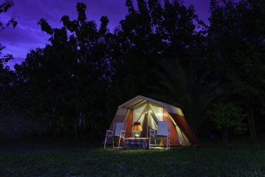 Geceleri kamp çadırları, antika kulübe çadırları, antika gaz lambaları, retro sandalyeler ve bir orman kamp alanındaki ahşap masalarda açık kahve yapma tesisleri olan bir grup kamp çadırı..
