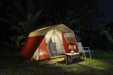 Geceleri kamp çadırları, antika kulübe çadırları, antika gaz lambaları, retro sandalyeler ve bir orman kamp alanındaki ahşap masalarda açık kahve yapma tesisleri olan bir grup kamp çadırı..