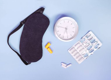 Uyuyan göz maskesi, çalar saat, kulak tıkaçları ve renkli arka planda izole edilmiş uyku hapları veya vitaminler.