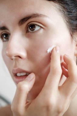 Yüzüne nemlendirici krem süren bir kız. Açık arkaplanda kusursuz derisi olan genç bir kızın fotoğrafı. Cilt bakımı ve güzellik kavramı
