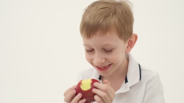 白いポロ不足しているいくつかの歯の小さな男の子は 光の背景に赤いリンゴを食べる スローモーション — ストック動画