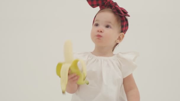 Kleines Mädchen Mit Kariertem Stirnband Und Rock Isst Große Banane — Stockvideo