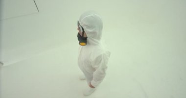 Beyaz kimyasal koruma giysisi giymiş ve gaz maskesi takmış bir adam kafasını yukarıdan çekim yapan kameraya çeviriyor. Kameraya doğru merdivenleri çıktı ve maskesini taktı.