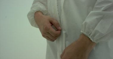 Siyah gözlüklü adam fermuarını çeker ve beyaz koruyucu elbisesini açar. Kameralar elleri takip ediyor, beyaz arkaplanda çekiliyor.