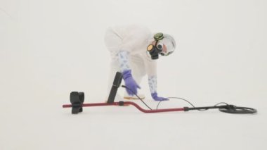 Beyaz kimyasal koruma giysisi, gaz maskesi ve yerden kalkan mor lastik eldivenli bir adam. Yerden metal detektörü alıyor ve düz duruyor. Beyaz arkaplanda çekilen.