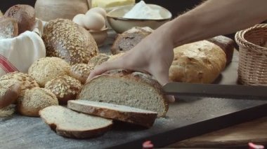 Erkek eller, çavdar ekmeği, susam tohumu veya yer fıstığı serpiştirilmiş buğday ekmeği de dahil olmak üzere diğer ekmeklerle dolu ahşap masanın üzerinde taze pişmiş Hollanda ekmeğini kesiyor. Hasır sepet yan tarafta duruyor.. 