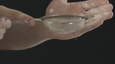 Erkek ellerinin küçük metal elek ile unu ellediği yakın plan. Siyah arkaplanda çekilen