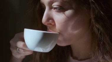 Uzun açık kahverengi saçlı ve uzun kirpikli bir kızın beyaz bir fincandan zevkle kahve içtiği yakın plan. Siyah bir arkaplanda yan yüz olarak çekilen