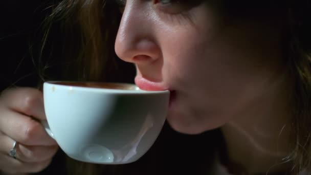 長いライトブラウンの髪と長いまつ毛が白いカップから喜びとコーヒーを飲んでいる女の子のクローズアップ 黒い背景の上で横に撮影 — ストック動画