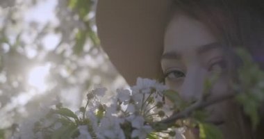 Gri gözlü, şapkalı ve elma ağacının çiçek açan dalını koklayan kahverengi tavşanlı kızın yakın çekimi. Açık havada çekilmiş. Bulanık arkaplan