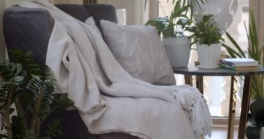 Koyu mor koltuk ve tencere ve kitapların olduğu yuvarlak kahve masası oturma odasının köşesine konur. Çiçek desenli beyaz yastık ve kalın kumaştan yapılmış püsküllü beyaz ekose koltukta.. 