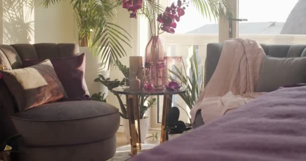 2つのアームチェアとラウンドコーヒーテーブル付きのベッドルームのコーナー フレームの前に紫色のカバー付きのベッドの一部 床やテーブルの上にはたくさんの開花装飾植物があります — ストック動画