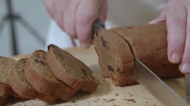 Baker (çerçevedeki erkek eller) taze fırınlanmış Hollanda üzümlü kekini ekmek bıçağıyla kurutulmuş kayısılarla kesiyor. Kırıntılar kesme tahtasına ve masaya düşmüş..