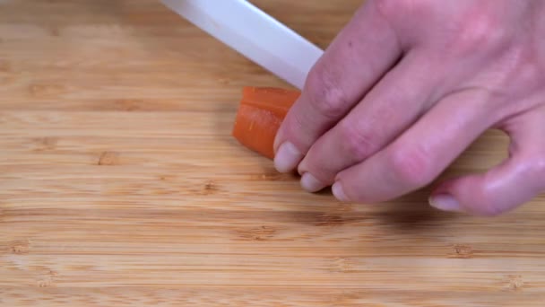 La mano femenina corta zanahoria con un cuchillo de cerámica blanca en una tabla de cortar de madera — Vídeo de stock