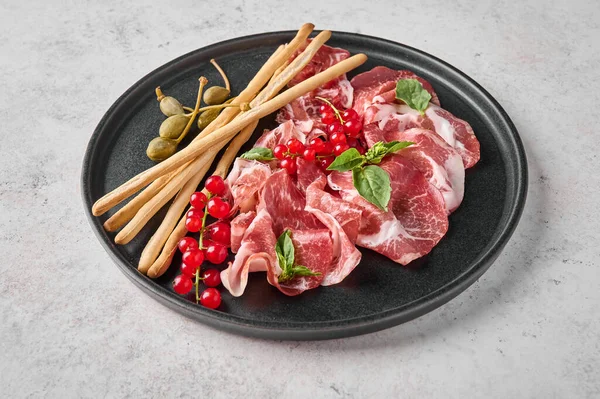 Close up prosciutto coppa di parma ham slices with basil, grissini sticks, capers and redcurrant — Stock Photo, Image