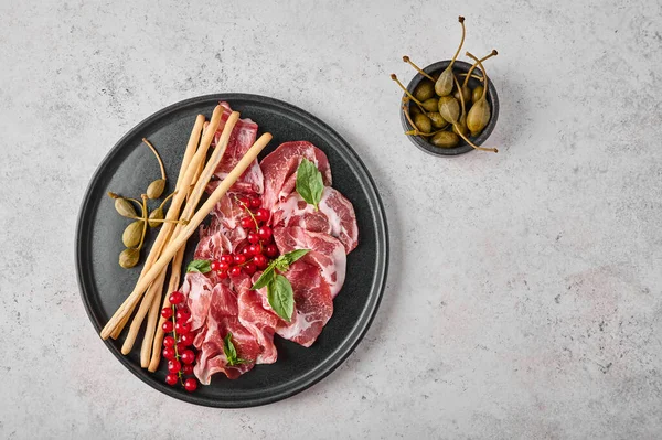 Slices of prosciutto coppa di parma ham with basil, grissini sticks, capers and redcurrant on dark plate — Stock Photo, Image