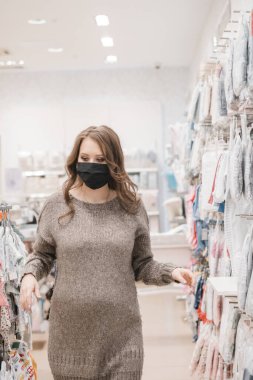 Hijyenik maskeli hamile bir kadın bebek kıyafetleri, bebek arabası ya da bebek arabası seçiyor. Müstakbel anne ve bebek alışverişi. Pandemik yaşam tarzına yönelik önleyici tedbirler.