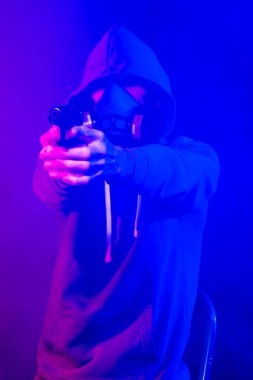 Bilinmeyen bir adamın portresi, karanlık arka planda neon maske takan bir hacker. Neon ışıklandırması yakınında silah tutan çift ırklı siber punk oyuncusunun seçici odak noktası