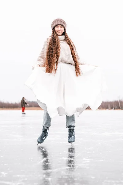 Woman ice skating outdoors on a pond. Outdoors lifestyle portrait of girl in figured skates. Smiling and enjoying wintertime. Wearing stylish down sweater, skirt, knitted mittens. Ready for skating