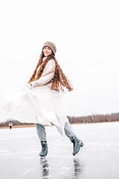 Woman ice skating outdoors on a pond. Outdoors lifestyle portrait of girl in figured skates. Smiling and enjoying wintertime. Wearing stylish down sweater, skirt, knitted mittens. Ready for skating