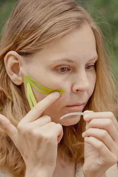 腎臓学の顔のテープを持つ美しい女性 女の子の美容師は 顔のキニーシオテープに顔を置きます 皮膚の概念を持ち上げる キネシオテーピング 女の子カット緑Kinesioテープとともにはさみ — ストック写真
