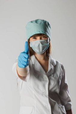 Tıbbi maskeli ve lateks eldivenli kadın başparmağını kaldırmış. Profesyonellerimizi seviyoruz. Koronavirüse karşı koruma. Tıbbi maske takan kadın doktor aşıyı teşvik ediyor