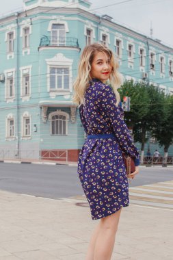 Bahar caddesinde yürüyen mavi elbiseli, yuvarlak çantalı güzel bir kadın. Şehir geçmişi. Şık bir turist kız hafta sonu gezisi boyunca şehirde gezmekten zevk alıyor..