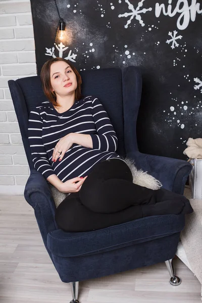 Beautiful pregnant girl with big belly near Christmas tree, having a rest on Christmas holidays before the new year. Happy new year concept. Pregnant woman at home. Expecting baby
