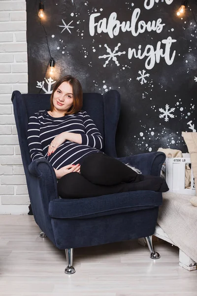 Beautiful pregnant girl with big belly near Christmas tree, having a rest on Christmas holidays before the new year. Happy new year concept. Pregnant woman at home. Expecting baby