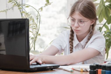 Akıllı liseli kız masasında oturmuş, kalemle not alıyor ve dizüstü bilgisayar kullanıyor. İnternette dil öğreniyor, evde okul ödevleri yapıyor, ders dinliyor.