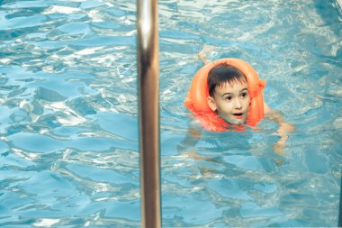 Yaz tatili gününde temiz suda krom tırabzanda duran, turuncu yüzen, havuz cankurtaranı cankurtaran yeleği giyen mutlu, tatlı çocuk. Mutlu çocuk için havuzda yaz aktiviteleri