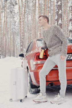 Gri elbiseli, kar üzerinde arabası olan, bavulu tutan ve çam ormanına giden genç ve modaya uygun bir adamın portresi. Kış tatili konsepti. Kışın dışarıda. Kayak merkezi, montaj merkezi