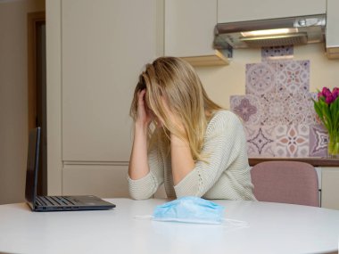 Evde dizüstü bilgisayarla çalışan güzel bir genç kadın. Masada tıbbi mavi maske var. Mutfakta üzgün ve üzgün bir kız var. Sağlık ve antivirüs koruma kavramı
