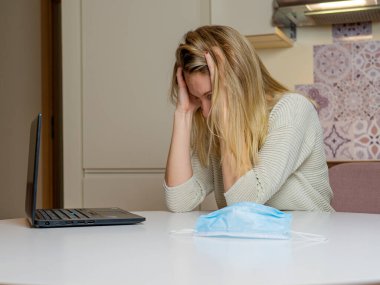 Evde dizüstü bilgisayarla çalışan güzel bir genç kadın. Masada tıbbi mavi maske var. Mutfakta üzgün ve üzgün bir kız var. Sağlık ve antivirüs koruma kavramı
