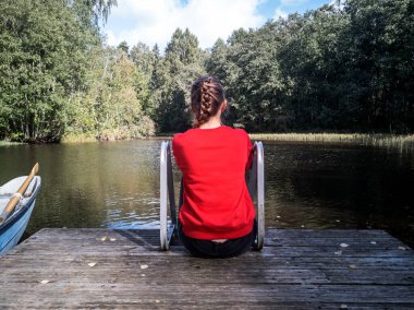 Nehir kıyısındaki ahşap iskelede oturan hayalperest kızın arka manzarası.