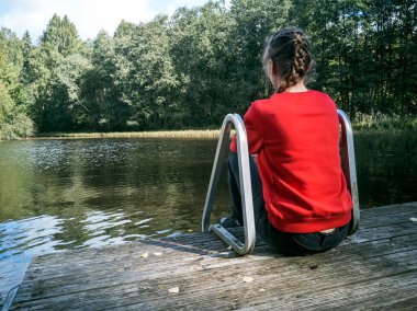 Nehir kıyısındaki ahşap iskelede oturan hayalperest kızın arka manzarası.