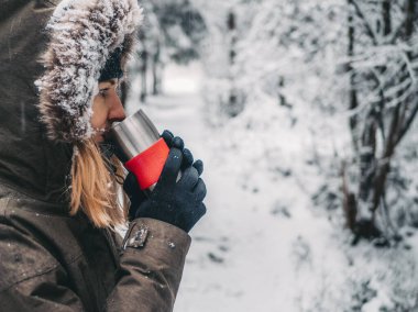 Genç bir kadın kışın sıcak içkinin tadını çıkarıyor. Arkaplanda kış boyunca karla kaplı ladin ağacı ormanı.