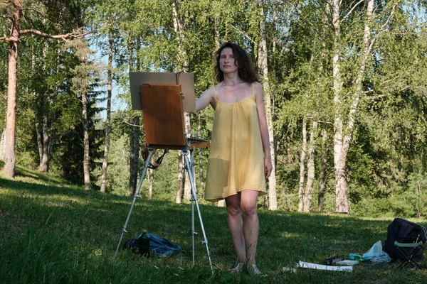 Girl in yellow dress paints a picture outdoors.