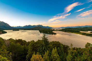 Panoramik bir manzara, Lago Moreno ve Lago Nahuel Huapi 'yi çevreleyen dağlık ufukta gün batımı gökyüzünü gösterir..
