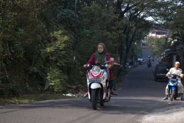 Malang, Endonezya 19 Nisan 2021: Malang 'da bir köy yolunda gün boyunca kask ve maske takmadan motosiklet süren bir kadın
