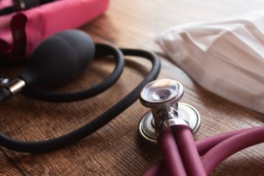 Pembe steteskop, solunumu ve kalp atışını inceleyecek ekipman, tansiyon ölçer ve koruma maskesi. Ahşap arkaplan.