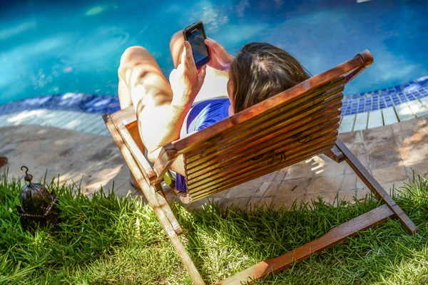 Молодая женщина отдыхает на шезлонге у бассейна и разговаривает по мобильному телефону — стоковое фото