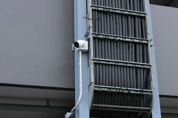 Dikey destek metal yapısına monte edilmiş bağlantı kutusuna bağlı bir güvenlik kamerası. Kamera, küçük siyah kravatlarla korunan çok sayıda kablo içeren bir kablo tepsisinin yanına yerleştirilmiş..