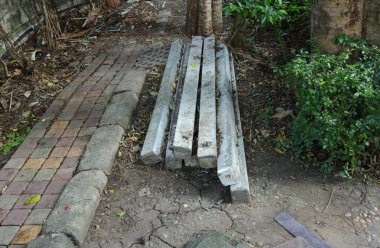 Bir yığın yıpranmış, dikdörtgen beton direkler bahçede kurumuş, çatlamış toprak üzerinde yatıyor. Kuru toprak, ağaç gövdesi ve yeşil çalı yapraklarıyla çevrili asfalt bir geçide yakın konumdalar..