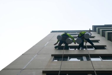 Tanımlanamayan iki işçi, halatlarla asılmış ve emniyet kemeri takmış, binanın dış tarafına dolgu macunu sürüyor. Aşağıdaki perspektif, parlak ve bulutlu gökyüzüne karşı işlerinin yüksekliğini ve zorlu doğasını vurgular..