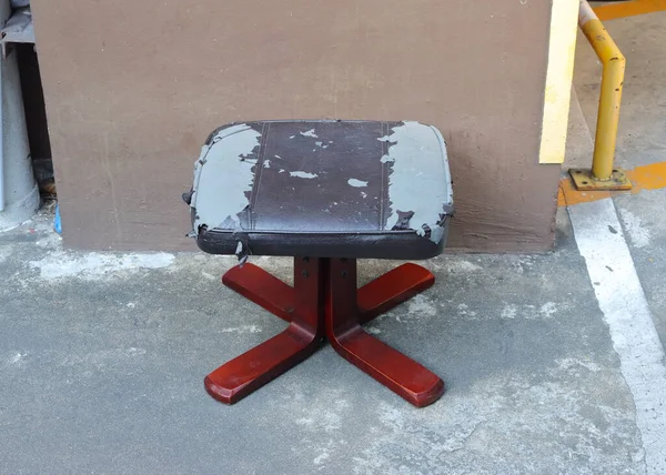 Açık havada gri beton bir yüzeye yerleştirilmiş bir tabure, kahverengi bir duvarın yanında. Dışkının dolgusu kötü durumda, koyu kahverengi sahte deri örtüsünün nerede soyulduğunu gösteriyor, altındaki gri maddeyi ortaya çıkarıyor.