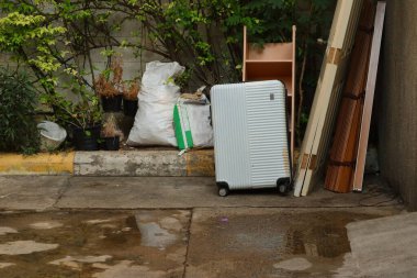 Islak beton bir yüzeyde, üzerinde dikey çıkıntılar olan yuvarlanan beyaz kabuklu bir bavul duruyor. Bavulda aşınma ve kir izleri var. Özellikle de alt ve yan kenarlarında sarımsı kahverengi lekeler..