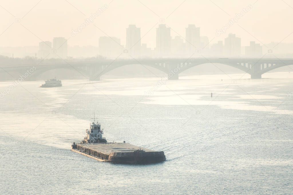 Barge and tugboat are on the river against the background of the bridge and urban views in the morning haze.