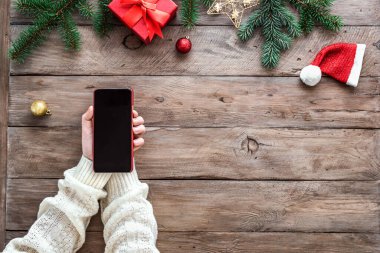 Noel süsleri, çam dalları ve Noel hediye kutusu olan ahşap arka planda akıllı cep telefonu tutan eller. Akıllı telefonlu Noel konsepti.