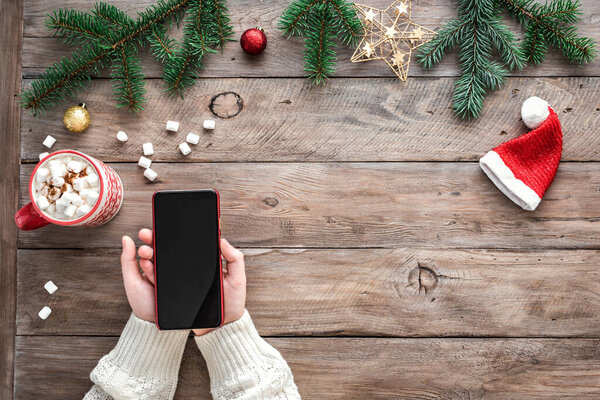 Hands holding smart mobile phone on wooden background with Christmas decor, pine branches and winter hot drink. Christmas concept with smartphone.